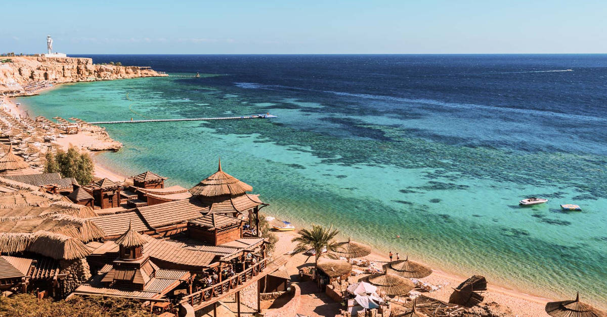 A scenic coastal view featuring a turquoise sea, sandy beach with straw umbrellas, and wooden buildings functioning as Egypt hotels, under a clear sky.